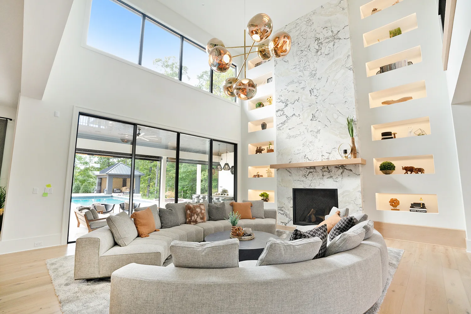 Alpha Fine Homes living room with a double-height marble fireplace, expansive windows and pool views